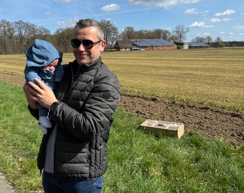 Marcus Möller hält ein Baby im blauen Kapuzenanzug im Arm. Dahinter liegen ein Feld und ein Hof, im Gras steht eine Kiste.