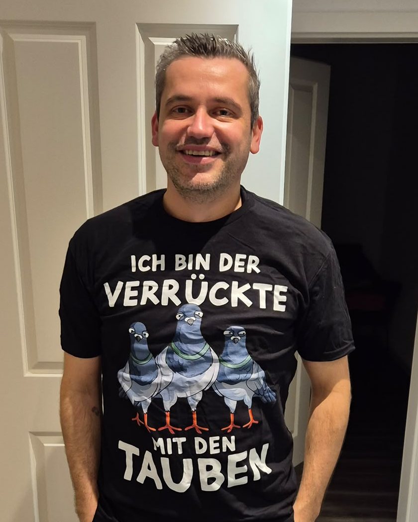 Marcus Möller lächelt in die Kamera. Auf dem schwarzen Shirt stehen drei Tauben und der Schriftzug „Ich bin der Verrückte mit den Tauben“.